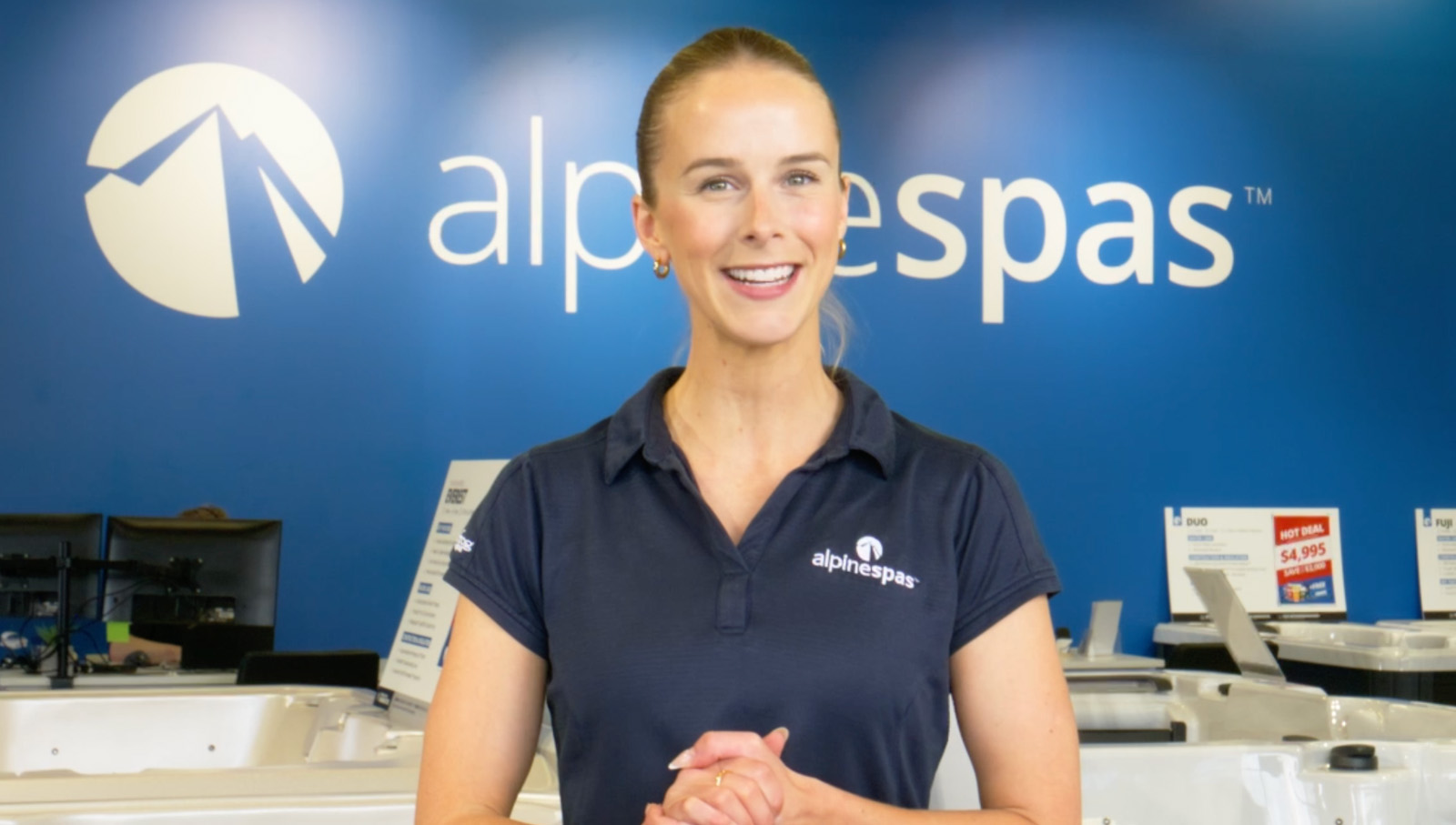 A woman in an Alpine Spas uniform stands smiling in a showroom with a blue Alpine Spas sign in the background.
