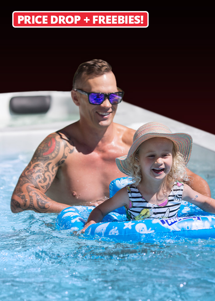A man and a young girl in a sun hat are enjoying a swim spa, with the girl sitting in a blue inflatable ring and a "Price Drop + Freebies!" banner above.