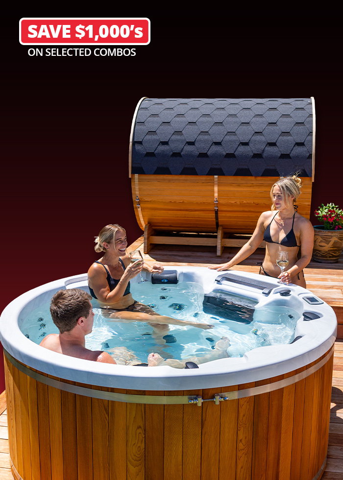 Two women and one man relax in a round outdoor spa with wooden panelling, holding drinks, with a barrel-shaped sauna in the background.