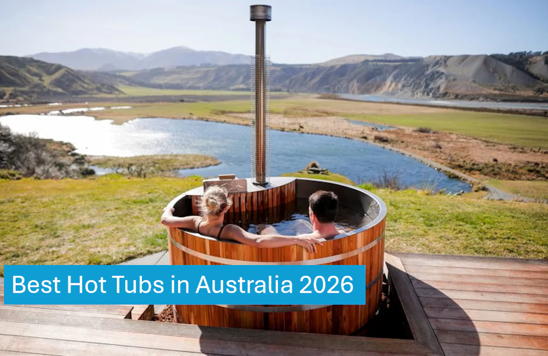 Two people relax in a round wooden outdoor tub overlooking a scenic river and mountains.