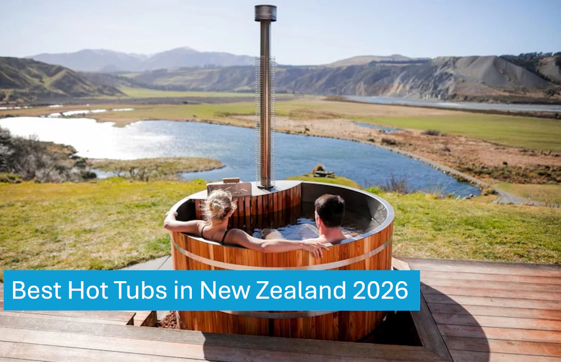 Two people relax in a wooden hot tub on a deck overlooking a scenic river and mountain landscape.