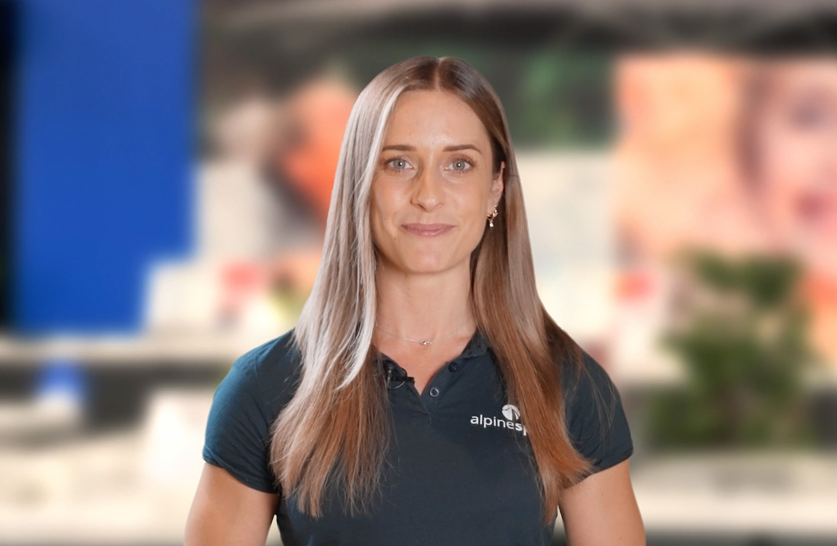 Woman with long straight hair wearing a dark polo shirt with the "alpines" logo, standing indoors with a blurred background.