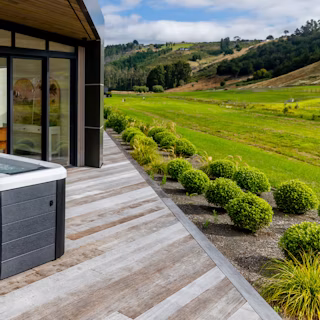 Four people relaxing in a hot tub on a wooden deck outside a modern house, with large windows and a scenic view of green hills and landscaped bushes.