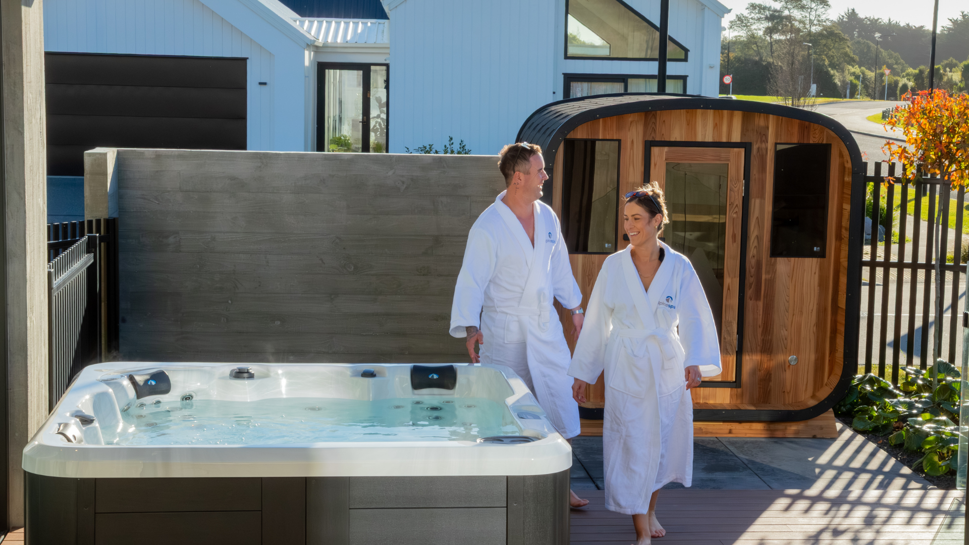 A man and woman in white bathrobes walk beside a hot tub and a wooden sauna on a sunny outdoor patio.