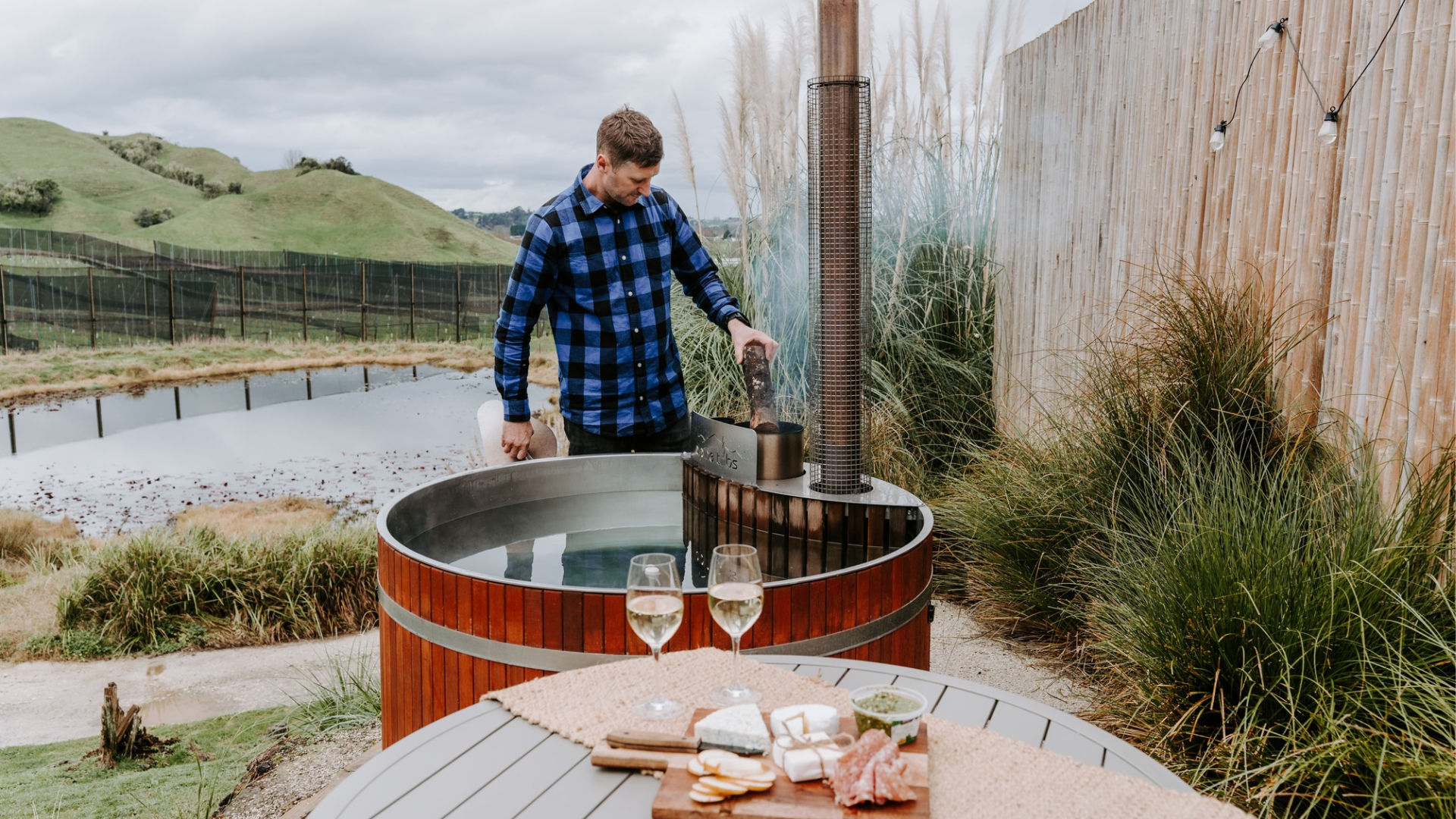 A man in a blue plaid shirt adds wood to an outdoor hot tub heater beside a pond, with a table in the foreground holding two glasses of white wine, cheese, crackers, and charcuterie.