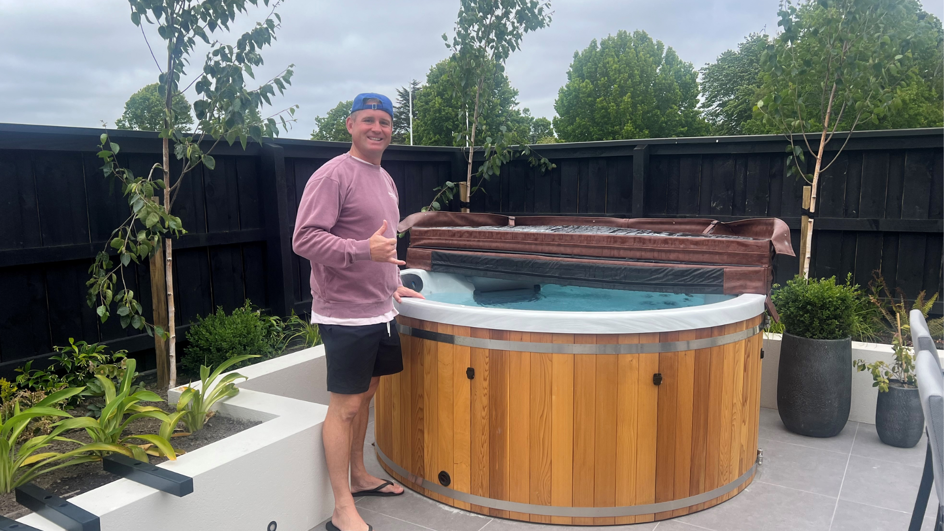 A man wearing a pink sweatshirt, black shorts, and a backwards blue cap stands beside a round wooden hot tub in a modern backyard, giving a thumbs-up gesture. The area is landscaped with small trees, plants, and a black wooden fence.