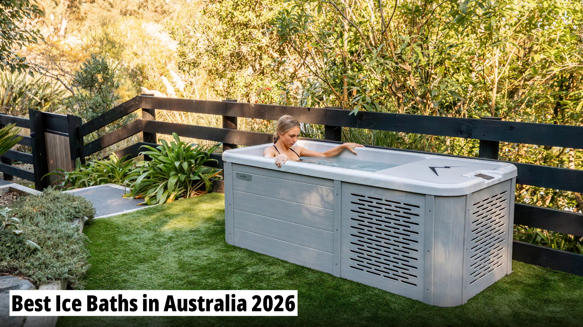 A woman is sitting in a modern outdoor ice bath in a garden setting with trees and a black wooden fence.