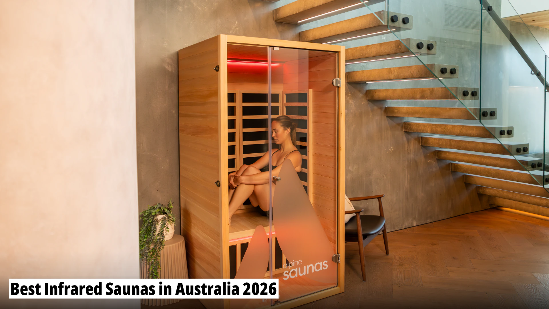 A woman sits relaxing inside a modern wooden infrared sauna cabin in a stylish home interior near a staircase.