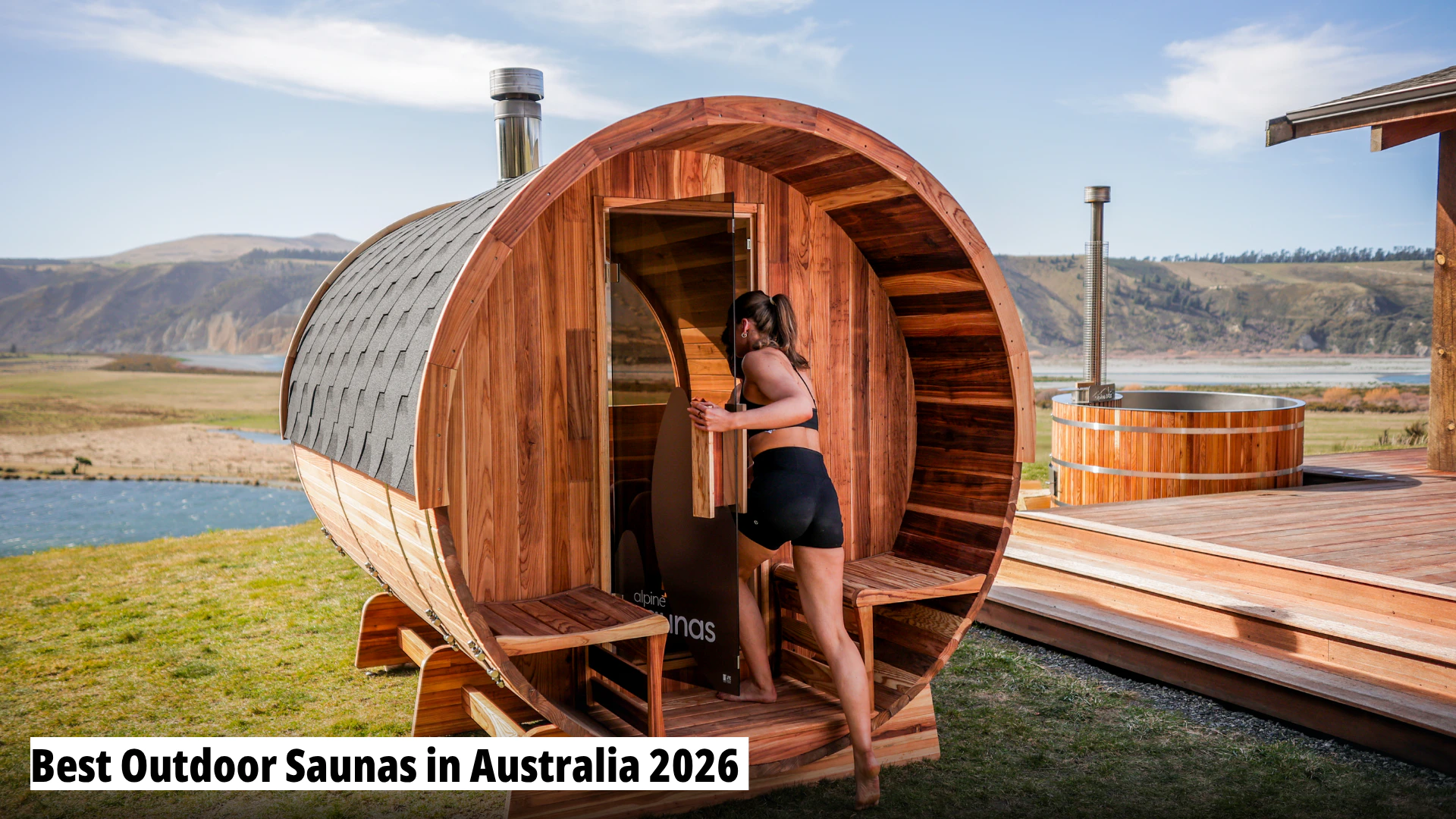 A woman in activewear enters a barrel-shaped wooden outdoor sauna set in a scenic landscape with a hot tub and lake in the background.