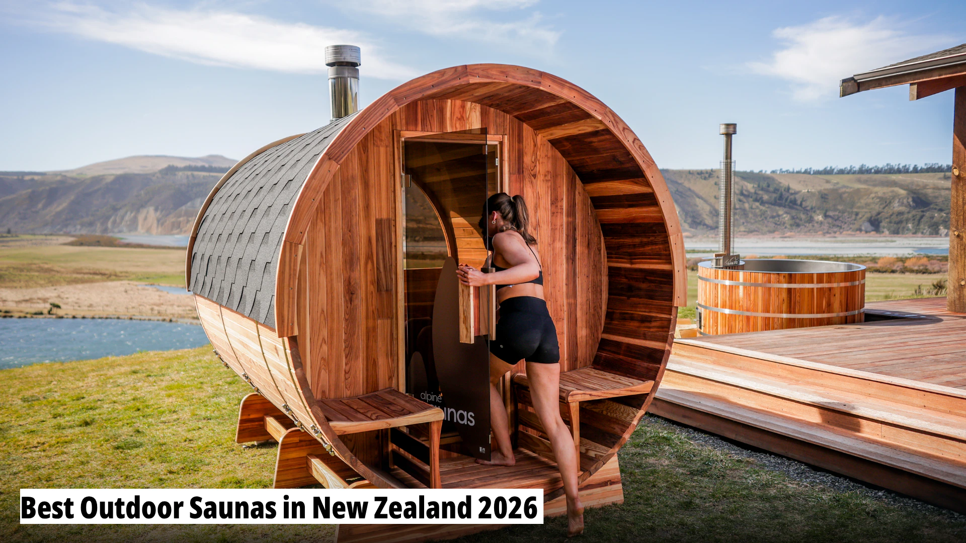 A woman in swimwear enters a wooden barrel-shaped outdoor sauna set on grass beside a deck, with hills and water in the background.