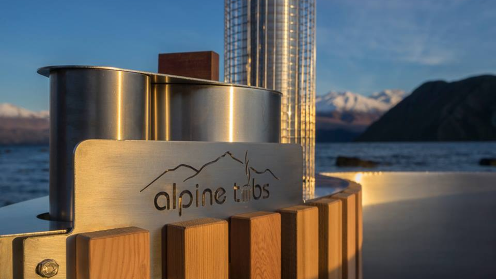 Close-up of a modern hot tub with a metal sign reading "alpine tubs," set outdoors by a lake with mountains in the background.