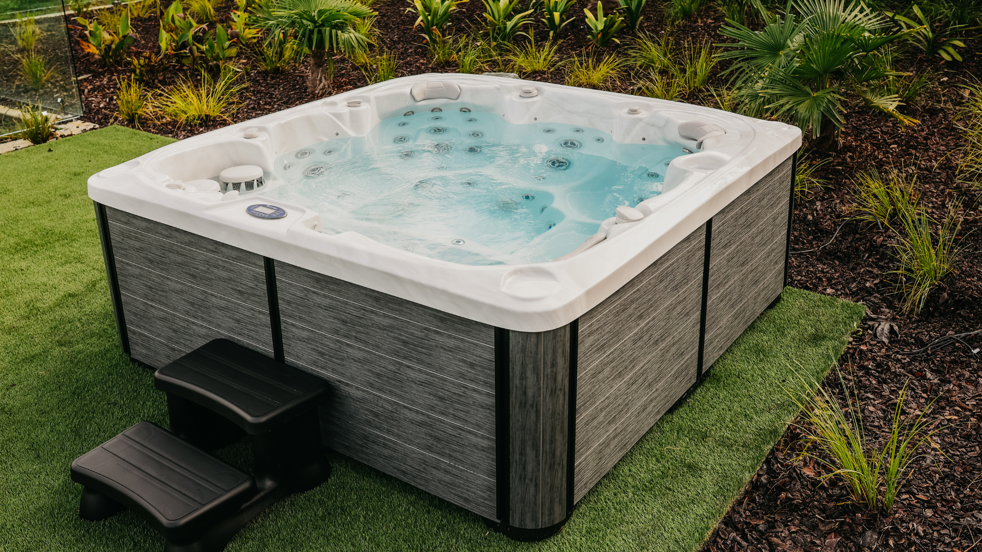 Outdoor square spa pool with bubbling water, gray panels, and black steps, surrounded by artificial grass and landscaped plants.