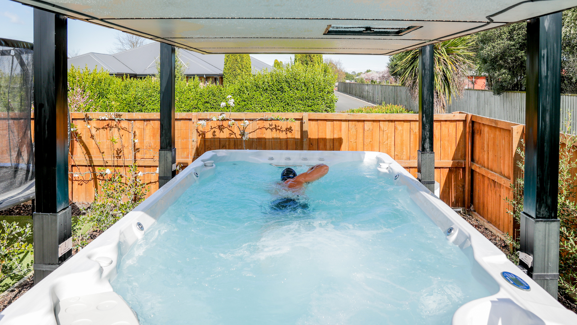 Person swimming in a backyard swim spa under a covered structure, with a wooden fence and greenery in the background.