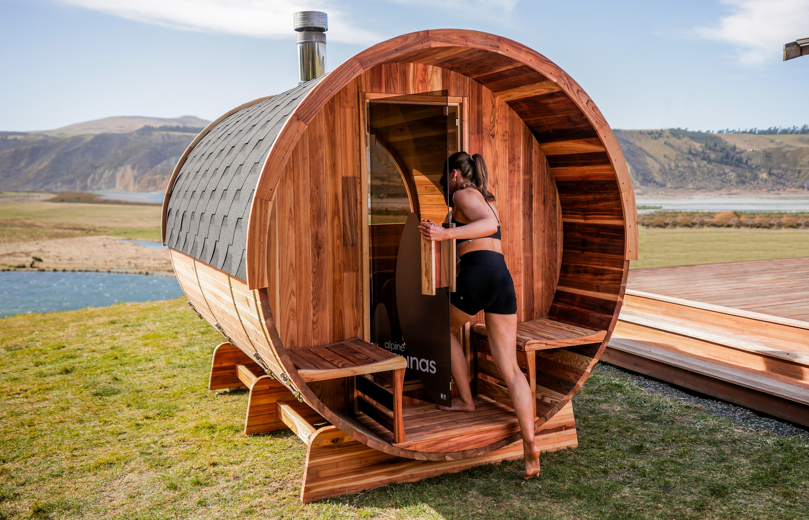 A person in athletic wear enters a barrel-shaped wooden sauna set outdoors on grass, with mountains and a river visible in the background.