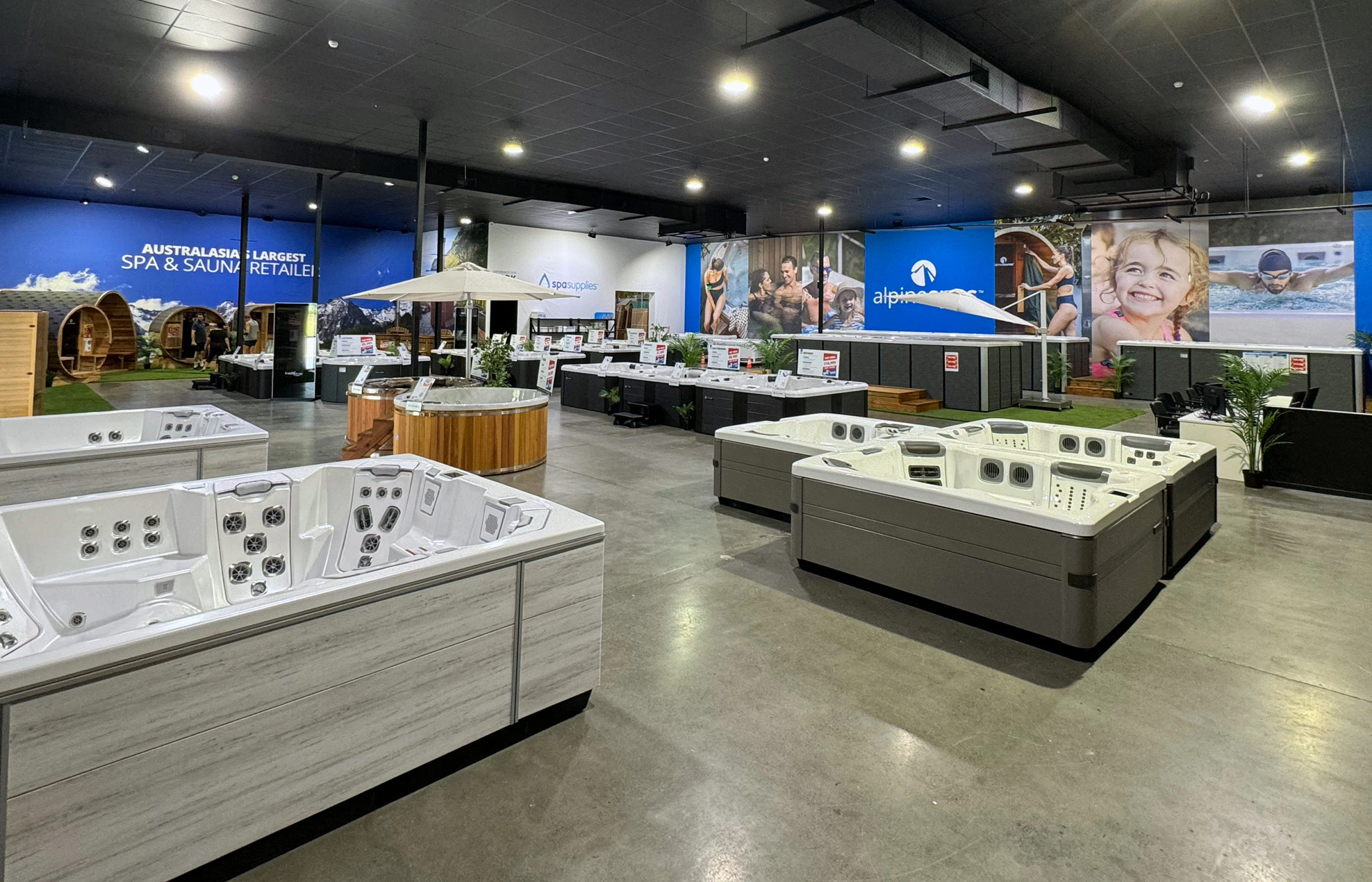 Spacious showroom displaying multiple hot tubs and saunas, with large promotional banners on the walls and spa accessories arranged throughout the area.