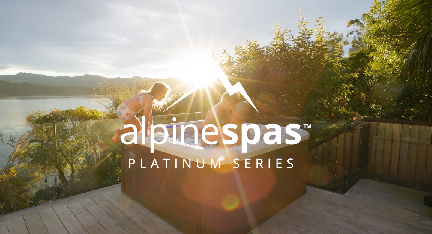 Two people relax in an outdoor spa on a wooden deck with sun shining through trees and mountains in the background.