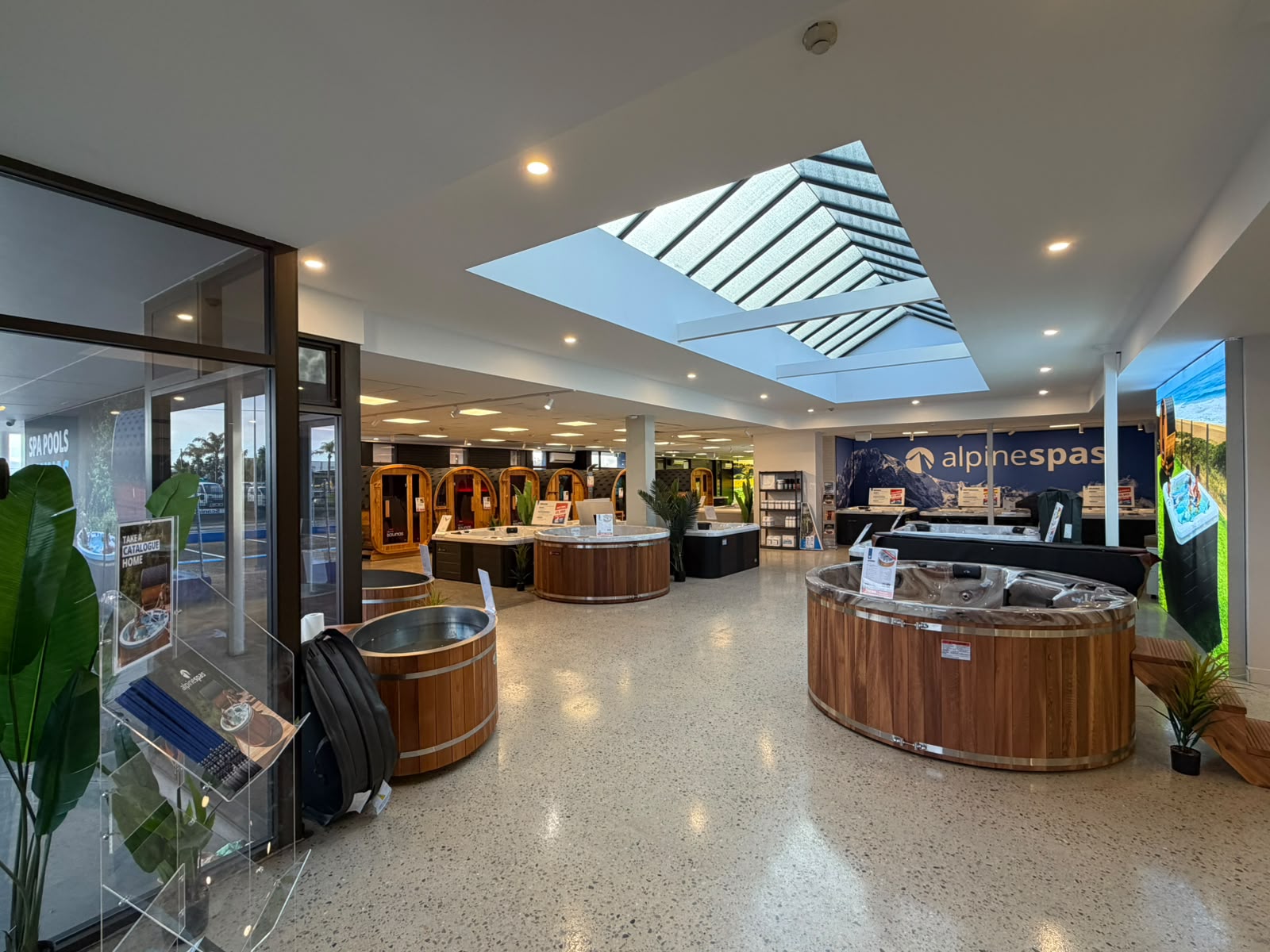 A showroom displays various wooden spa products, including circular tubs and barrel-shaped units, with bright lighting and signage for Alpine Spas.