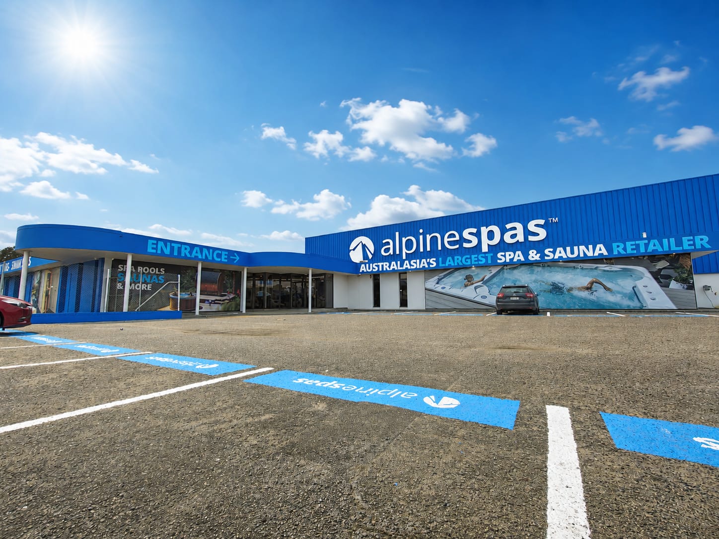 A large blue retail store with "alpine spas" branding, car park spaces, and signage advertising spa pools and saunas under a sunny sky.