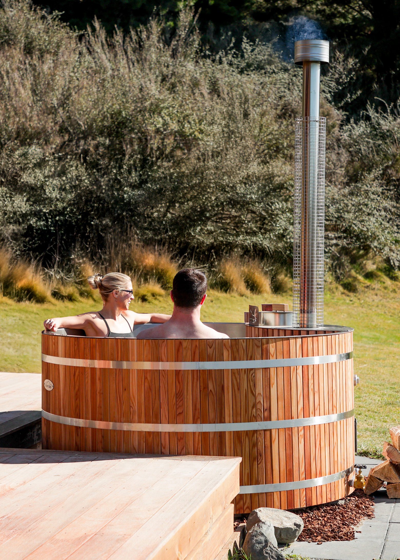 Two people relax in an outdoor wooden hot tub with a metal chimney, surrounded by greenery and natural landscape.