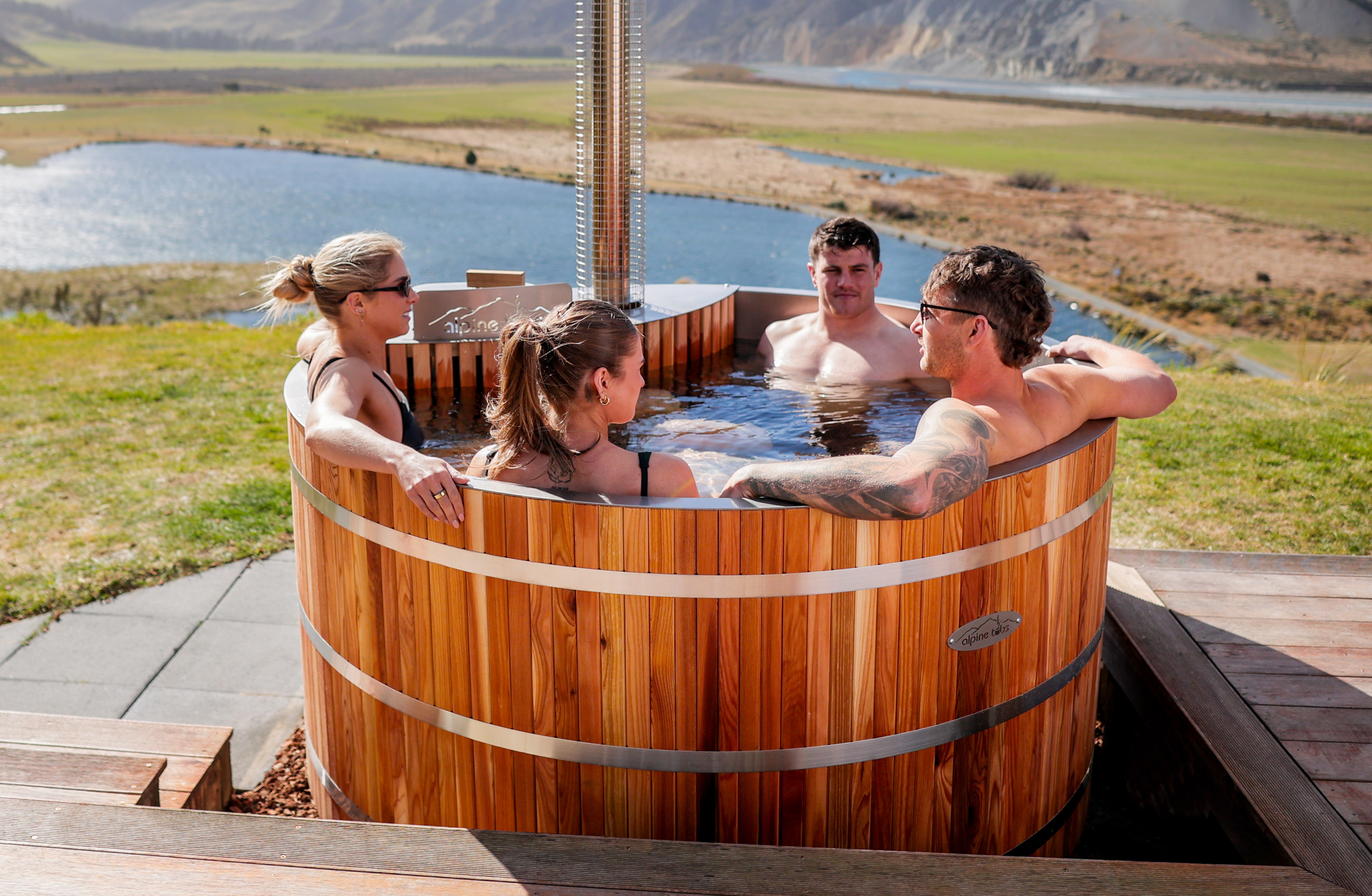 Four people relax in a wooden hot tub outdoors, with a scenic background of a lake, grassy field, and mountains.