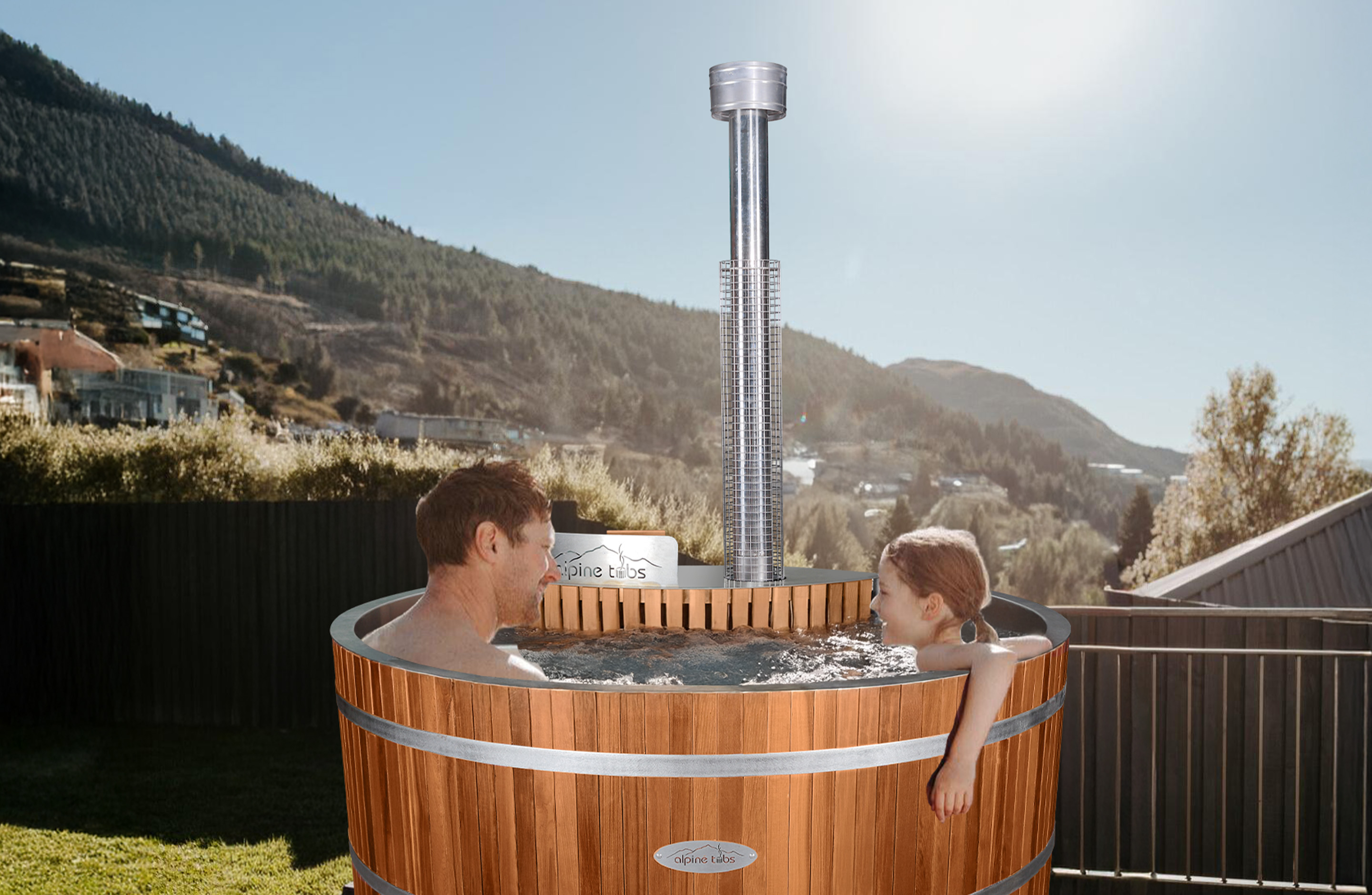 A man and a young girl relax in a round wooden hot tub outdoors, with a mountainous landscape and clear sky in the background.