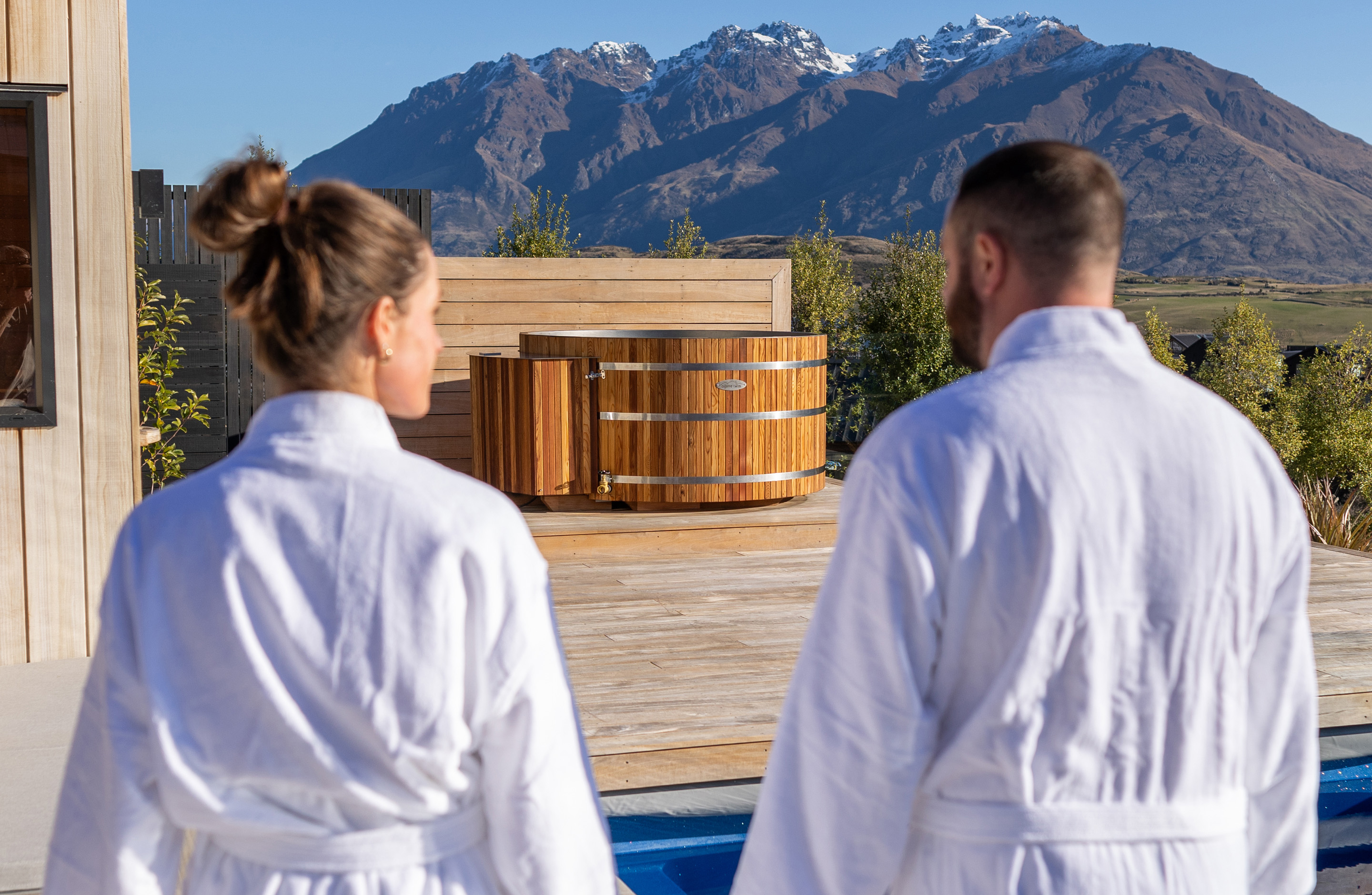 Two people in white robes stand on a wooden deck facing a wooden hot tub, with mountains and clear blue sky in the background.
