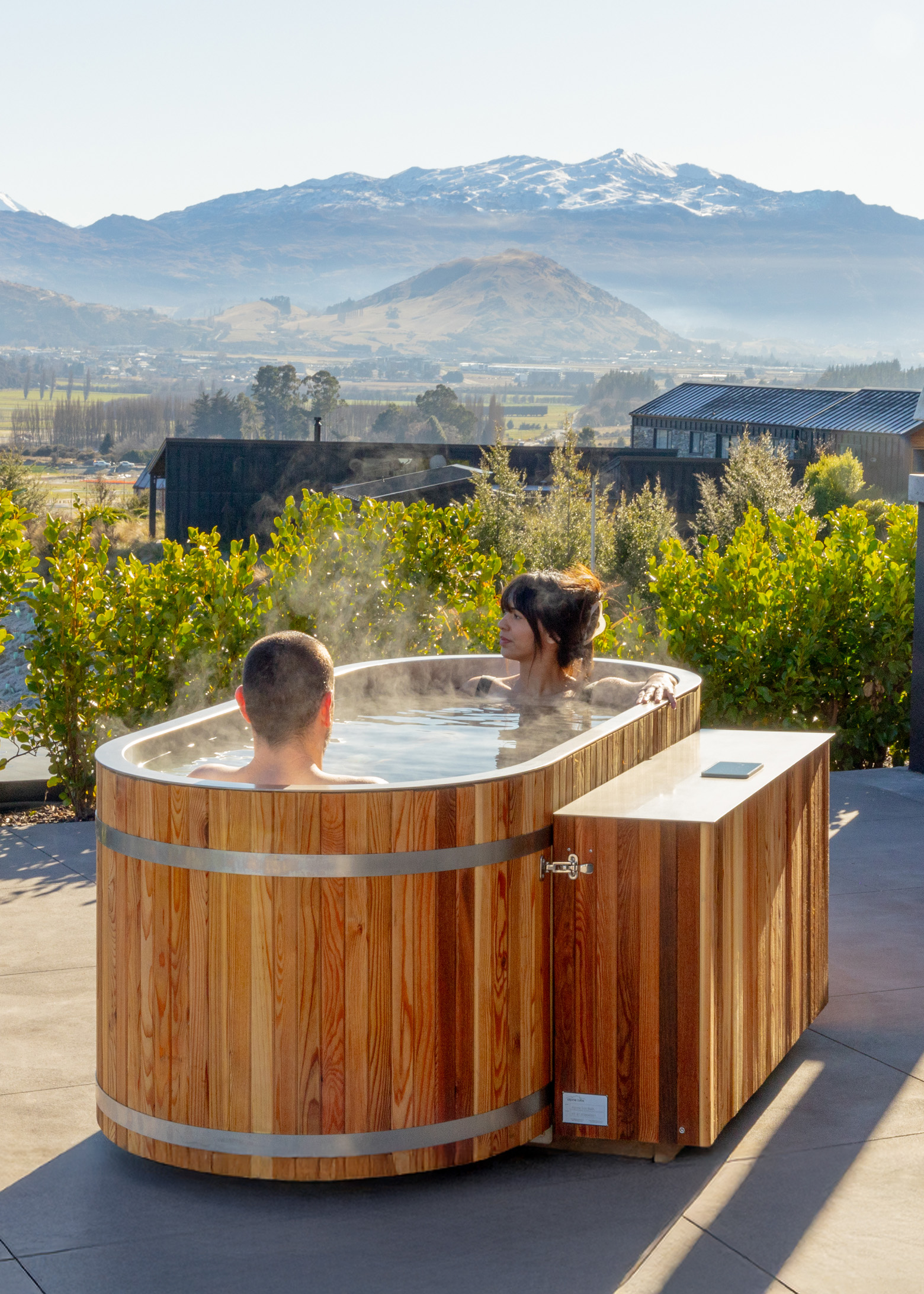 Two people relaxing in a steaming wooden hot tub outdoors, surrounded by greenery, with mountains and buildings visible in the background.