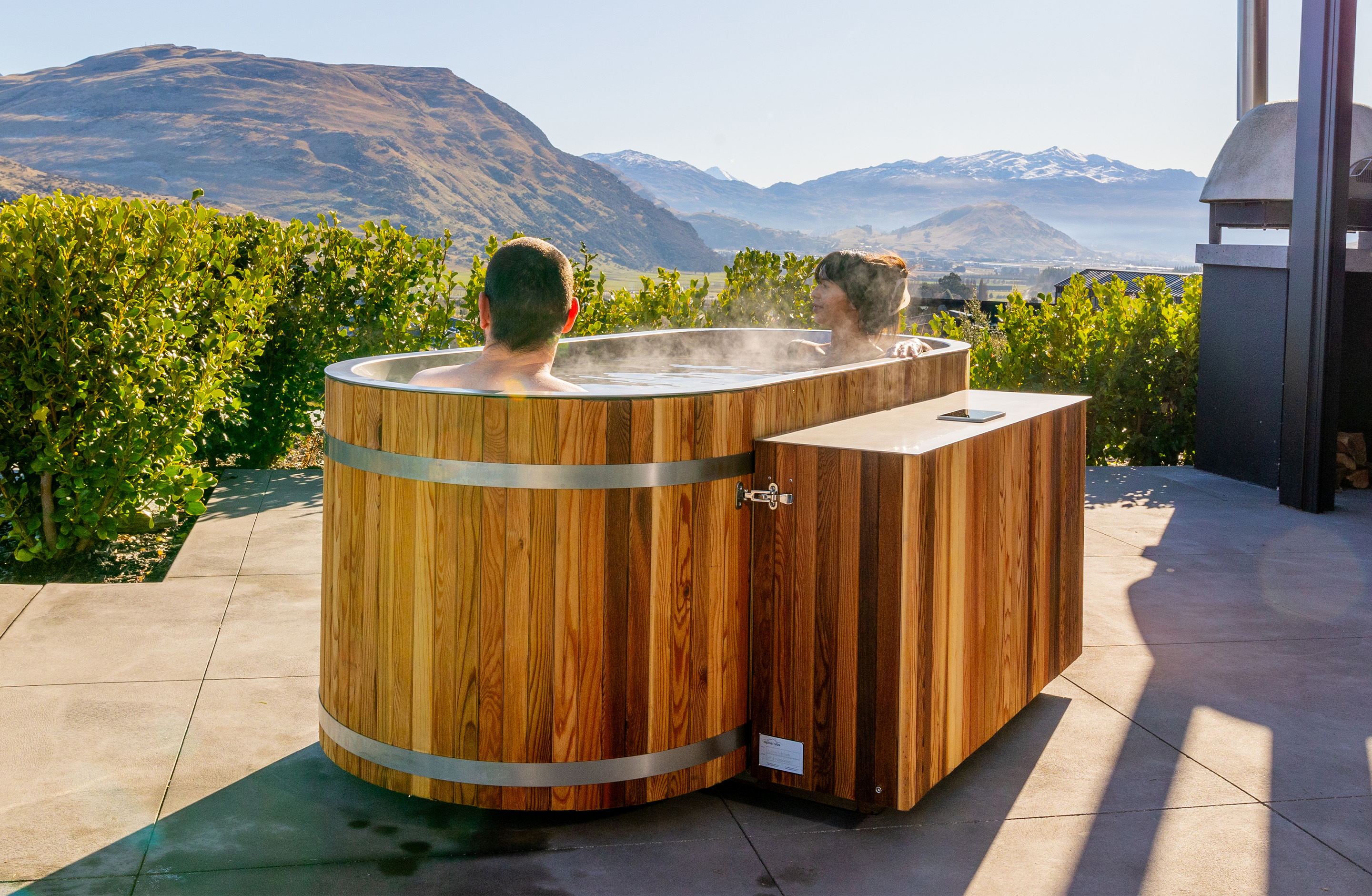 Two people relax in a steaming wooden hot tub on a patio, surrounded by green bushes, with mountains and a clear sky in the background.