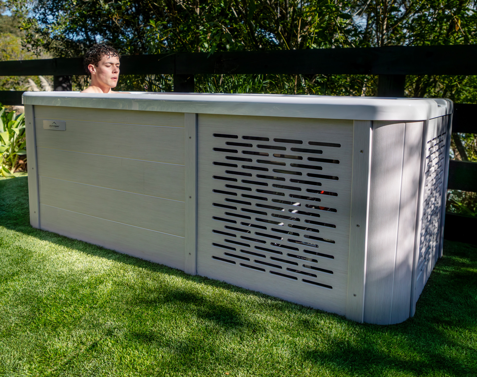 A person relaxes in an outdoor pool with a light-coloured cabinet and ventilation panel, set on artificial grass near a wooden fence.