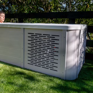 A person relaxes in an outdoor pool with a light-coloured cabinet and ventilation panel, set on artificial grass near a wooden fence.