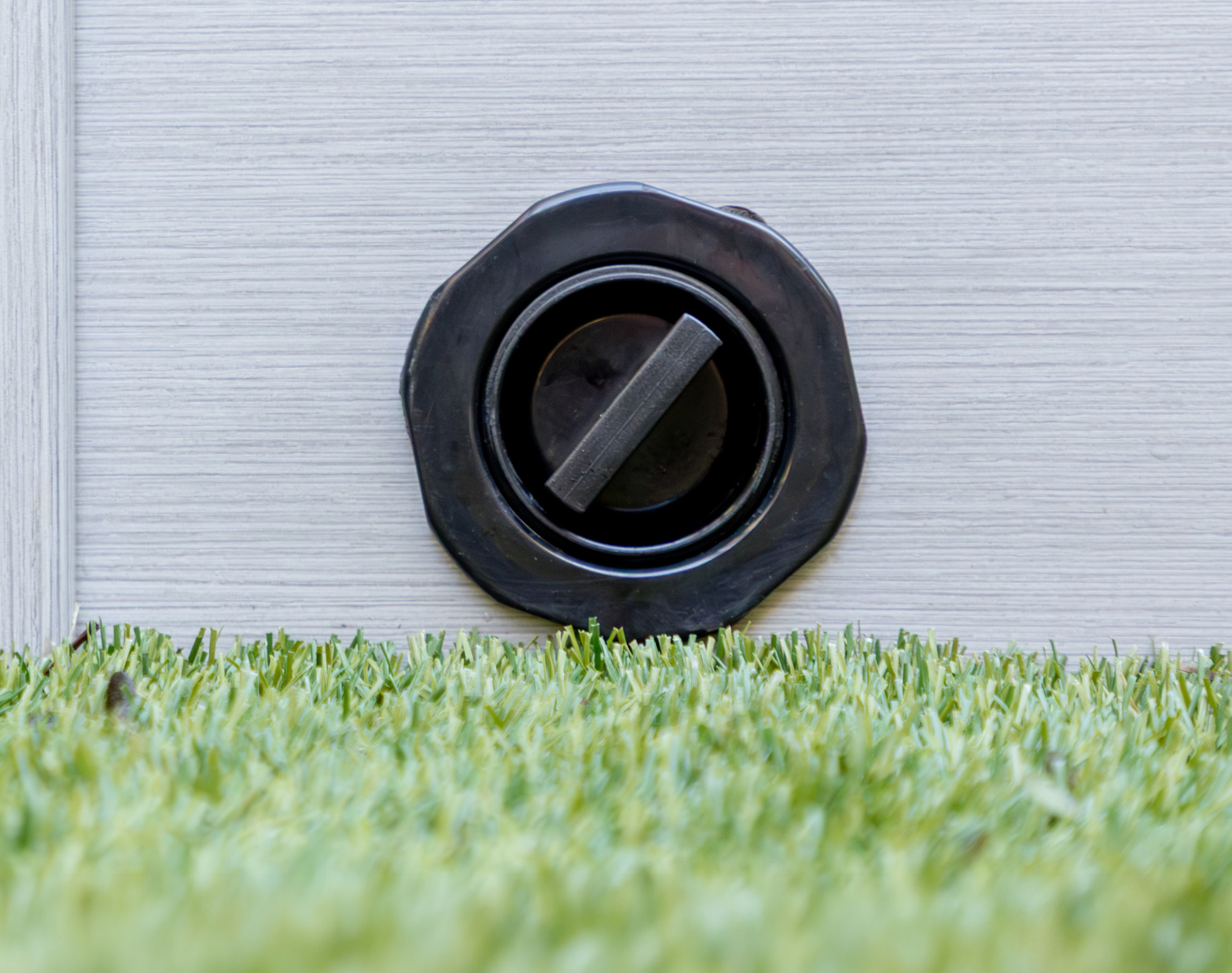 A black circular drain valve is mounted on a light-coloured panel above artificial grass.