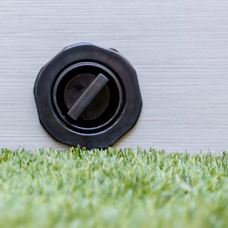 A black circular drain valve is mounted on a light-coloured panel above artificial grass.