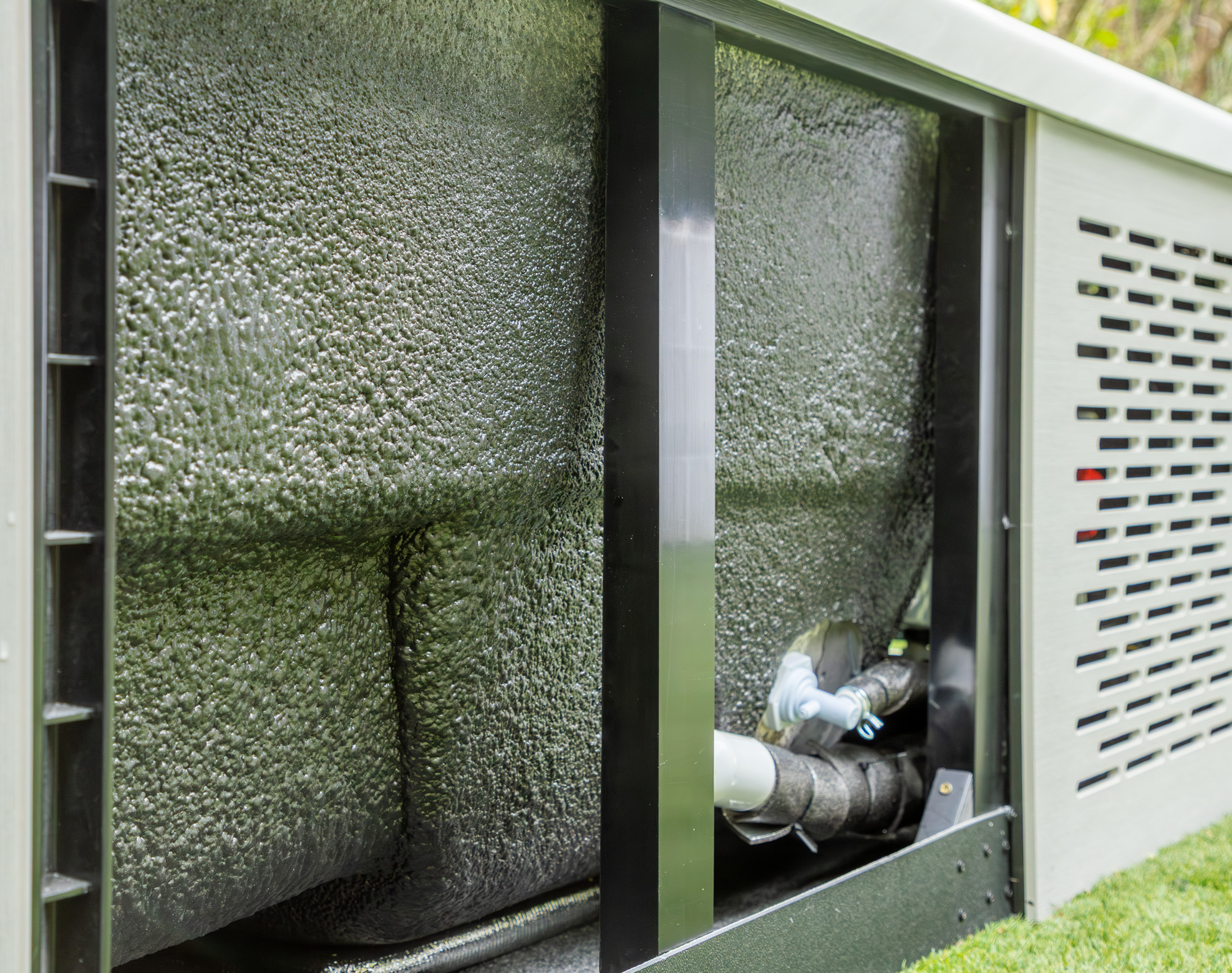 Close-up view of the insulated underside and plumbing of an outdoor unit with a textured black surface and metal frame, set on grass.