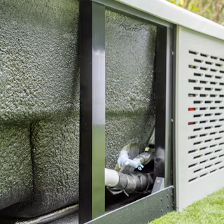 Close-up view of the insulated underside and plumbing of an outdoor unit with a textured black surface and metal frame, set on grass.
