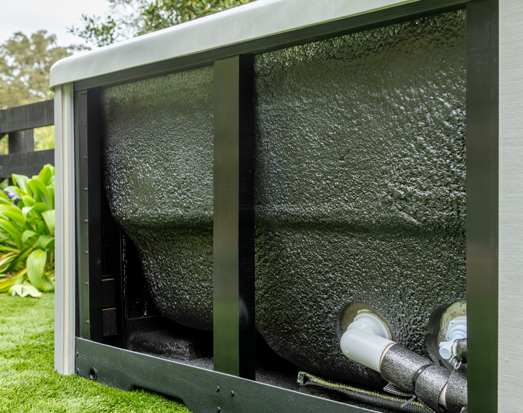 A close-up view of insulated piping and foam-covered outer wall on the side of an outdoor unit, with plants and grass visible nearby.