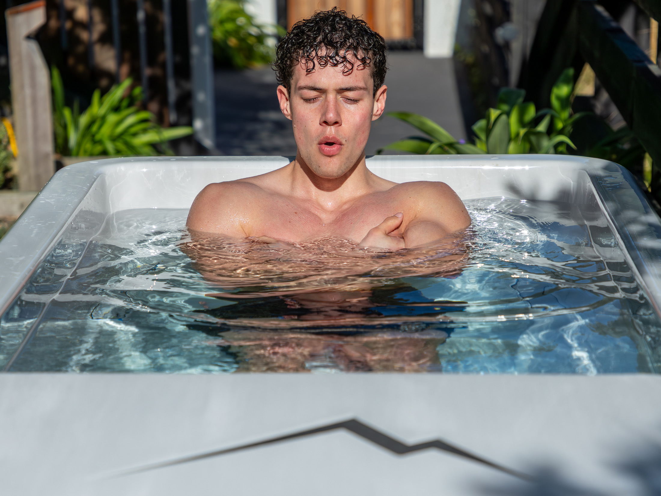 A man with curly hair is sitting in a white outdoor pool, arms crossed over his chest, eyes closed, and appearing to exhale deeply.