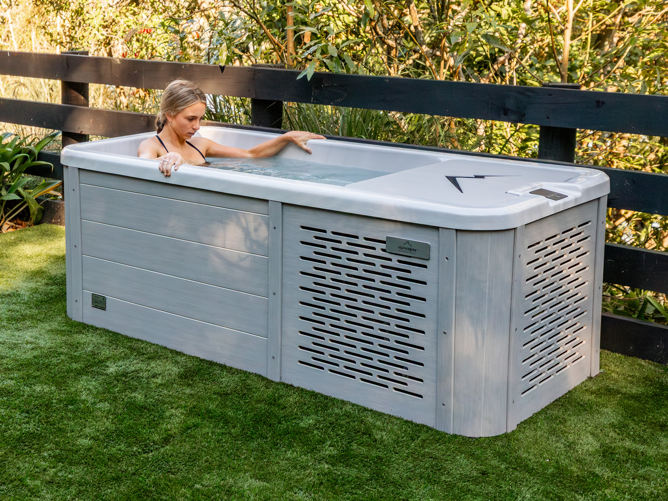 A woman relaxes in a rectangular outdoor spa pool with a light grey exterior, set on artificial grass near a black wooden fence and garden plants.