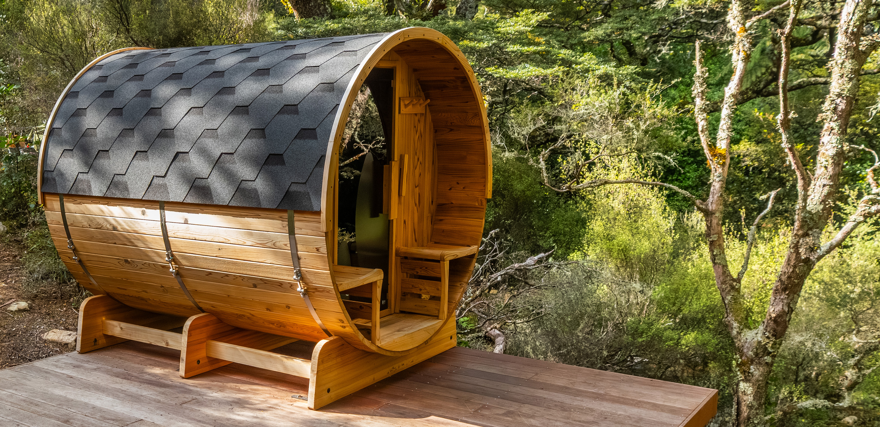 A barrel-shaped cedar outdoor sauna with a shingled roof sits on a wooden deck surrounded by lush forest.