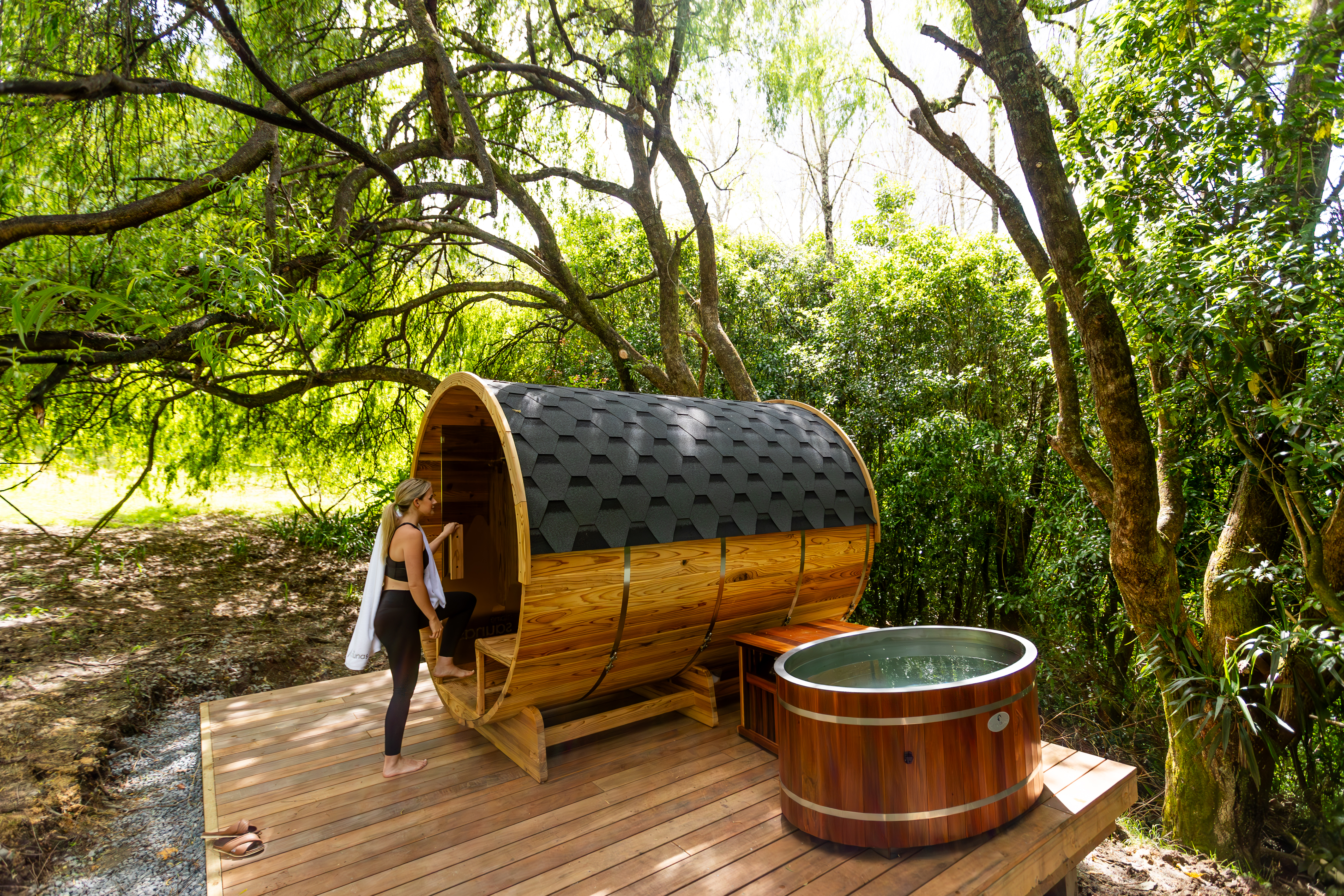 A person is stepping into a round wooden sauna next to a small round outdoor tub on a wooden deck surrounded by lush trees.