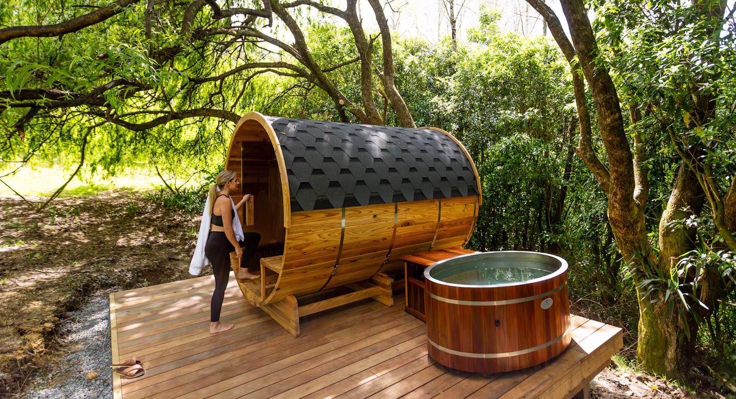 A person is stepping into a round wooden sauna next to a small round outdoor tub on a wooden deck surrounded by lush trees.