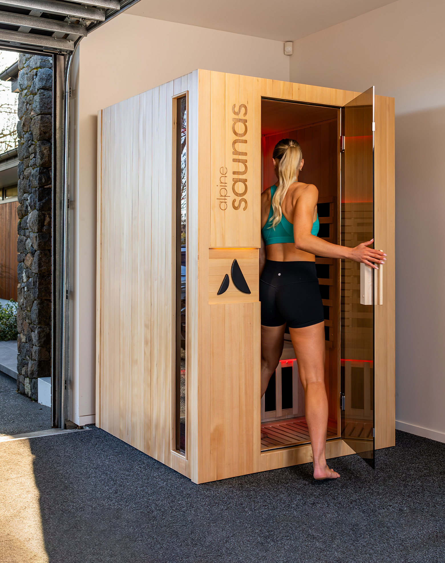 A woman in athletic wear is stepping into a wooden sauna cabin located inside a garage with the door open, letting in natural light.