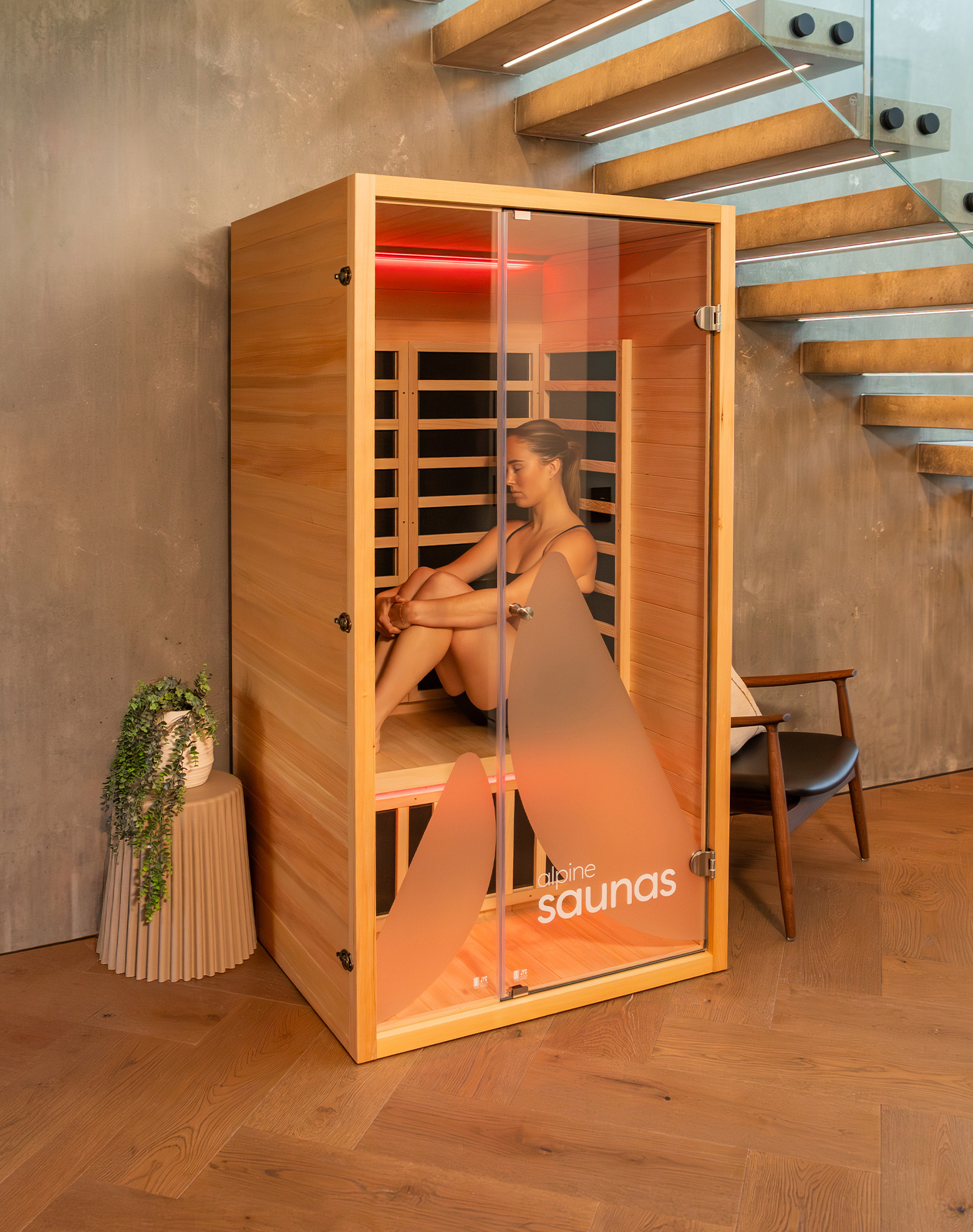 A compact wooden sauna with a glass door is placed under a modern staircase. A woman sits inside the sauna, and the words "alpine saunas" are printed on the door. A small side table with a potted plant and a chair are nearby.