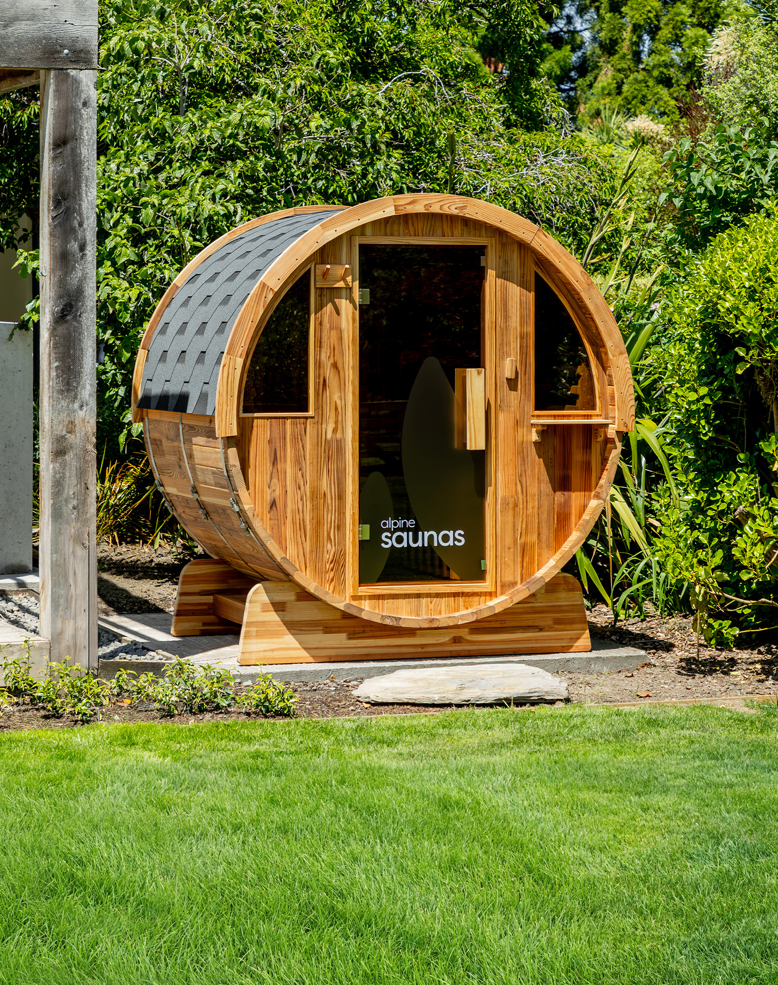 A barrel-shaped wooden sauna with a glass door and windows sits on a stone platform in a lush, green backyard surrounded by trees and bushes.