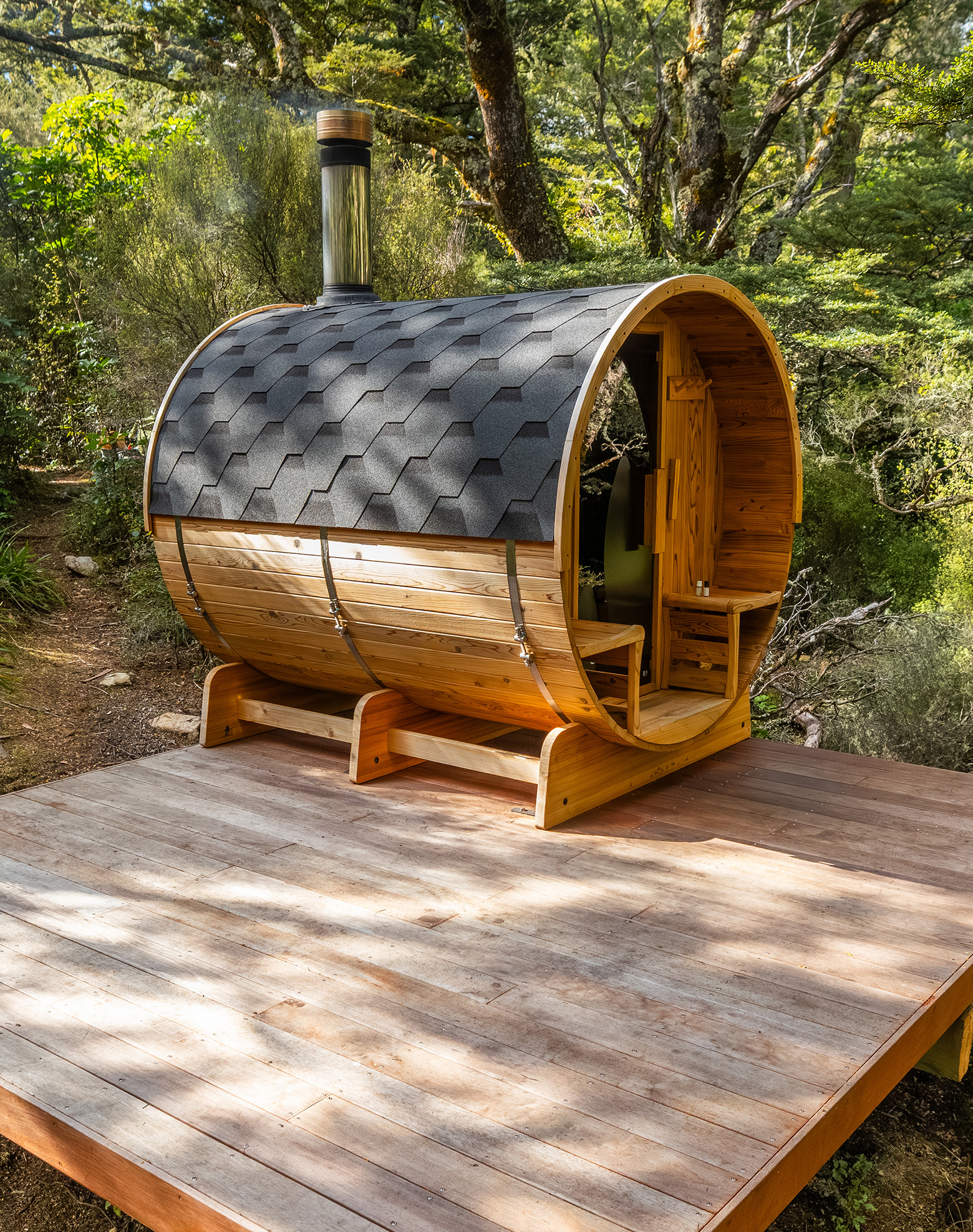 A small barrel-shaped wooden sauna with a shingled roof and metal chimney sits on a wooden deck surrounded by trees and greenery.