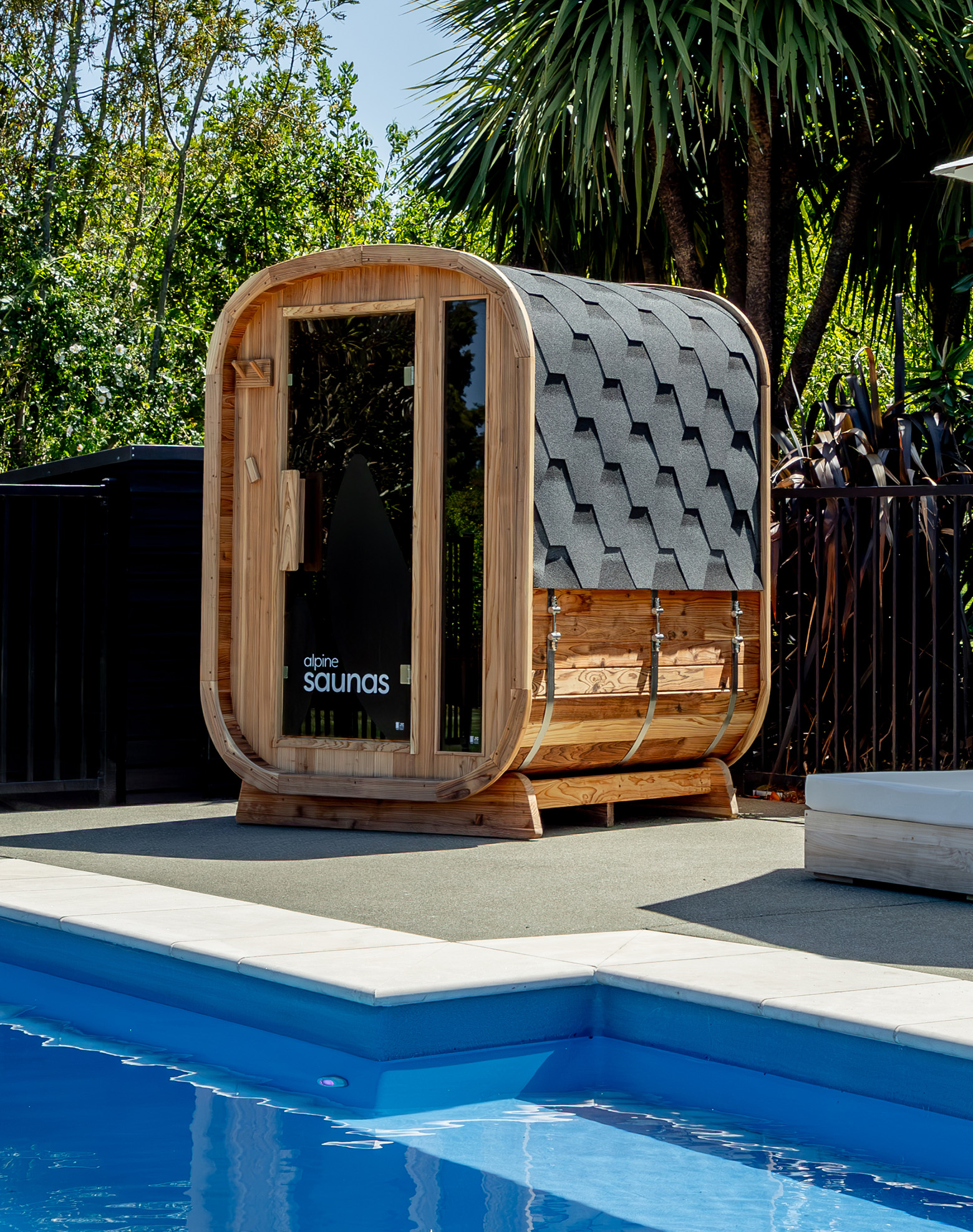 A small wooden barrel-shaped sauna with a shingled roof sits beside a blue outdoor swimming pool, surrounded by greenery and palm trees.