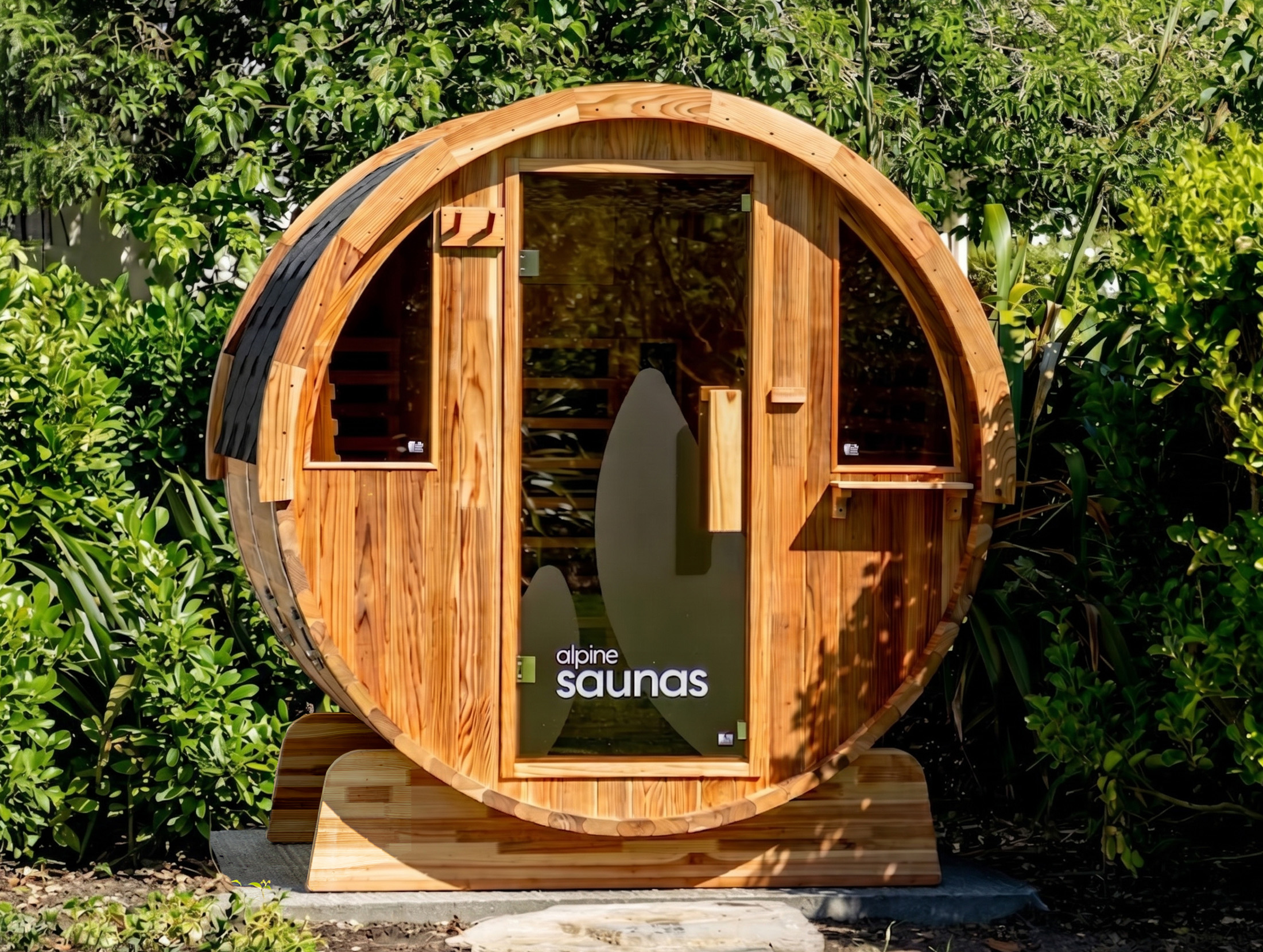 A round wooden outdoor sauna with a glass door sits surrounded by lush green plants.