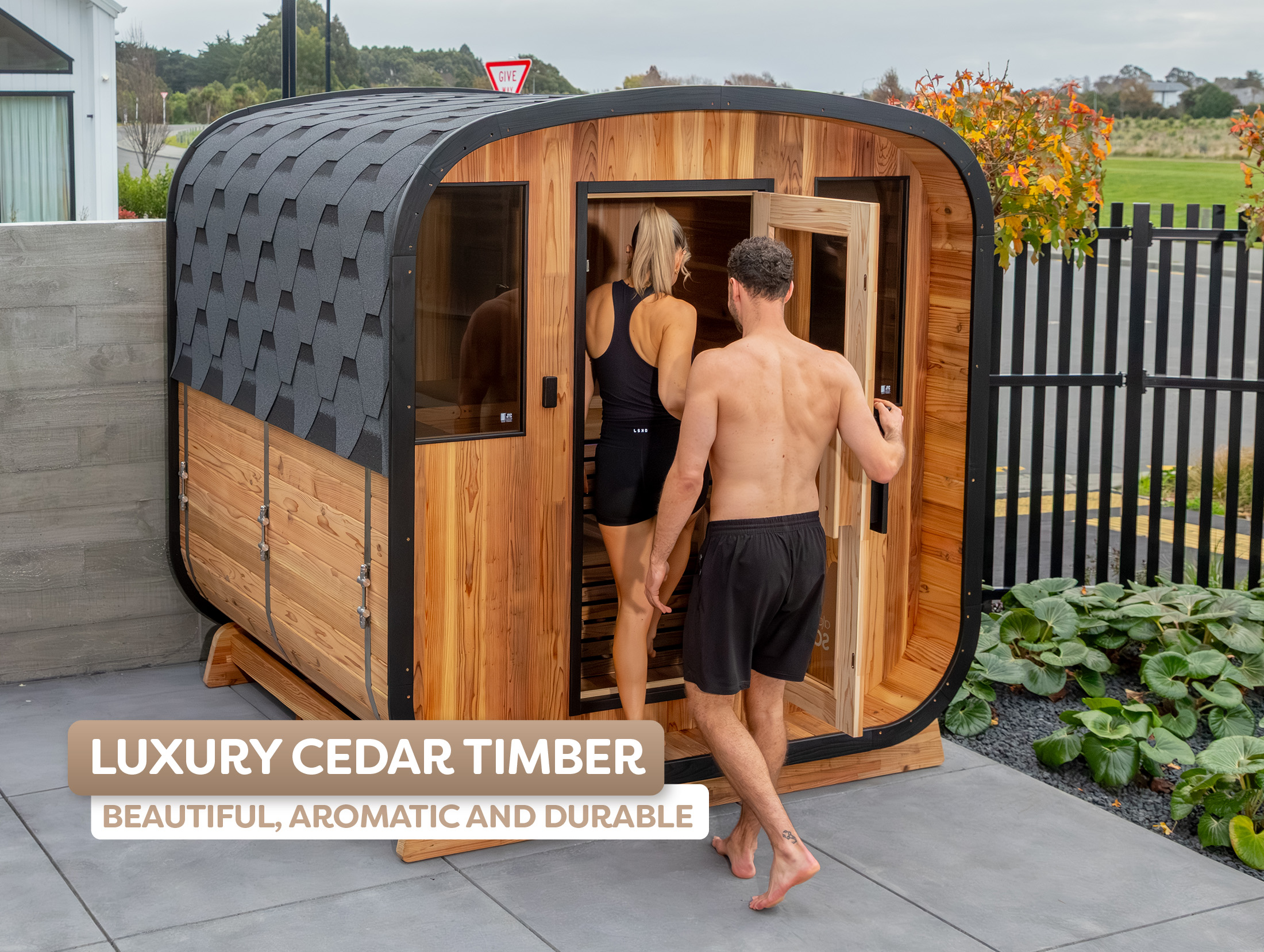 A man and woman in swimwear enter a modern cedar timber outdoor cabin with a curved roof, set on a patio beside a garden.
