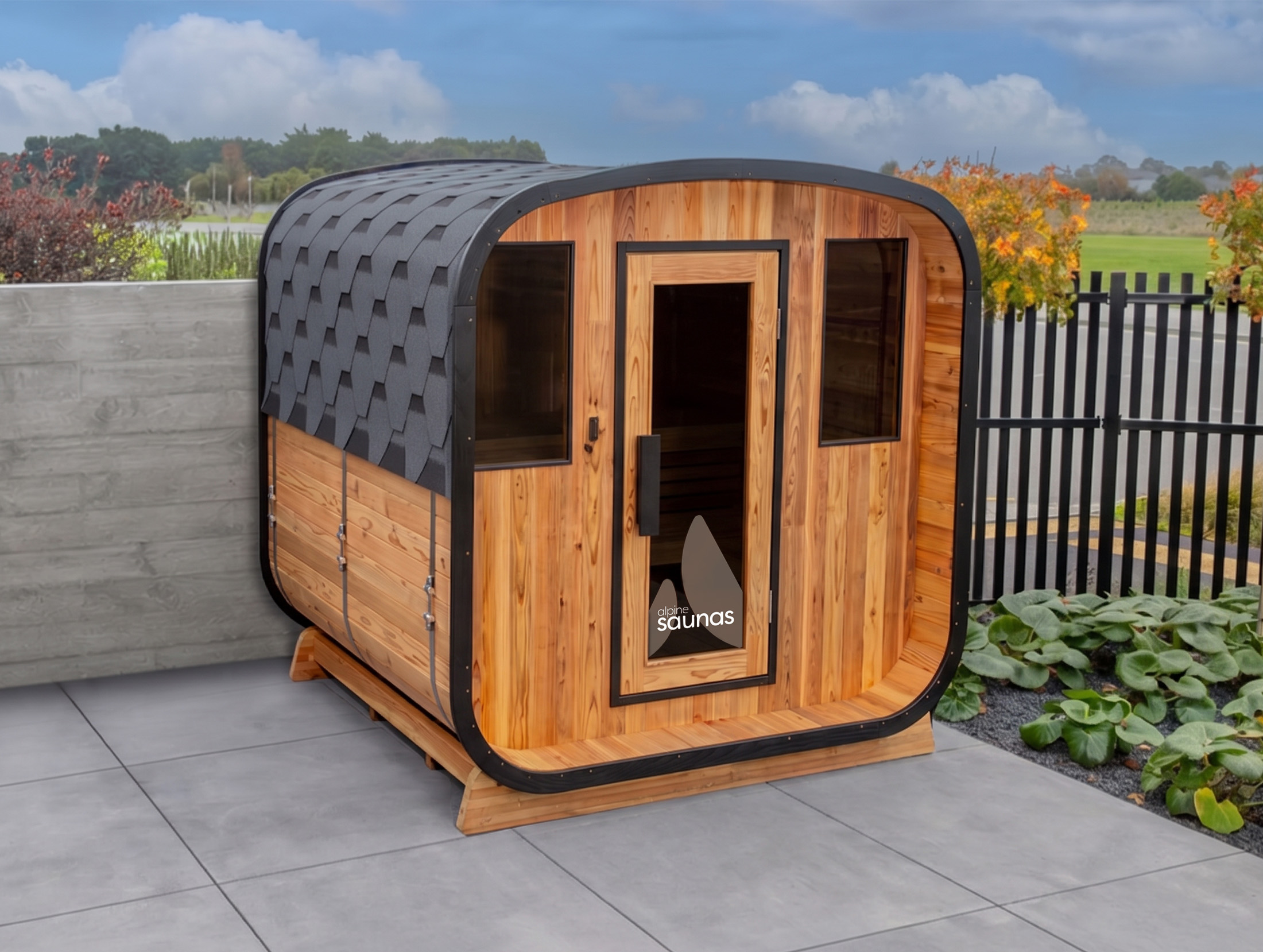 A modern wooden outdoor cabin with a shingled roof and large windows, set on a tiled patio beside a fence and garden.