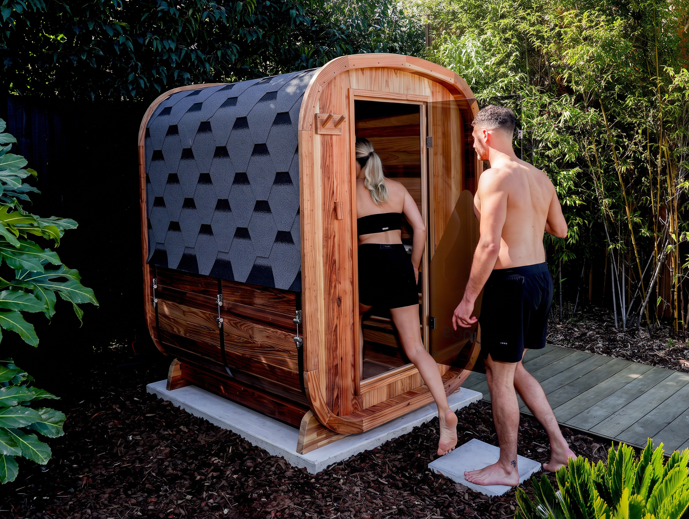 A woman and a man in swimwear are entering a small wooden outdoor sauna surrounded by greenery.