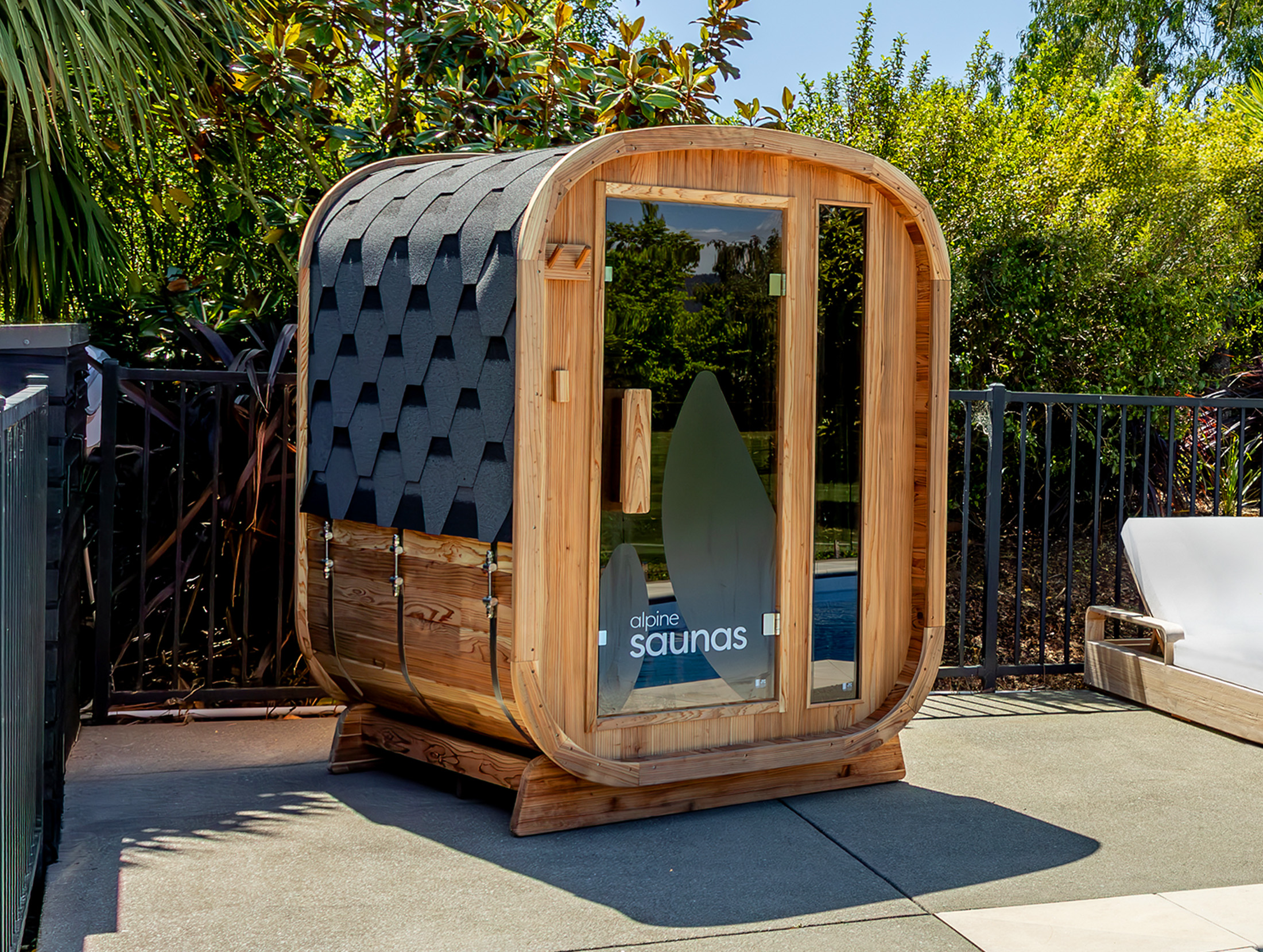 A modern wooden sauna with a curved roof and glass door is positioned outdoors on a patio near greenery and a sun lounger.